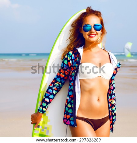 Outdoor lifestyle portrait of surfer girl posing at California beach, have perfect sportive tanned body, having active time at lonely exotic island, bright outfit, sunny colors, holding surf board.