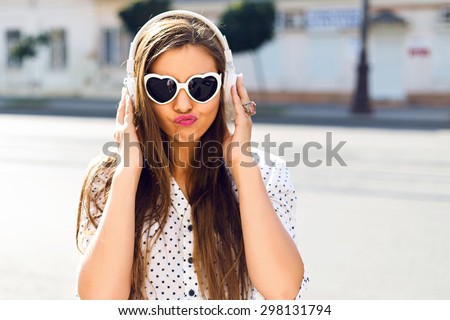 Close up funny portrait of sexy young pretty girl listening music on big earphones, wearing cute sunglasses, posing at city center at sunny summer day.