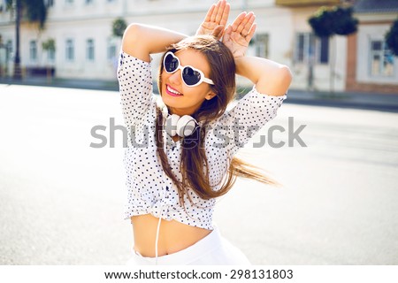 Young pretty hipster girl having fun, going crazy, imitating bunny ears by her hands, big amazing smile, fashion vintage outfit, sunglasses and headphones, posing at city center europe, cute emotions.