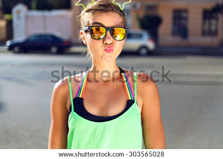 Young pretty hipster girl making funny grimaces and kiss, wearing bright summer clothes, mirrored sunglasses and funny hair accessory with neon stars.