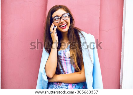 Close up lifestyle portrait of cute smiling brunette girl with amazing long hairs, hipster glasses and bright lips, speaking with her friends by her smart phone , positive emotions, wall background.
