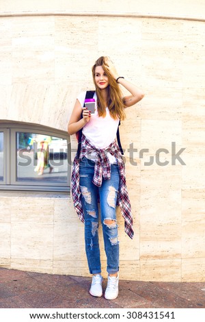 Lifestyle summer portrait of pretty blonde hipster woman, wearing street fashion casual outfit, denim and plaid shirt, blonde hairs and bright make up, drinking take away coffee, travel with backpack.