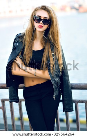 Fashion portrait of stunning sexy blonde woman, wearing brutal total black outfit, sunglasses, leather jacket, long hairs, vintage toned film colors, windy cold weather, autumn time, urban style.
