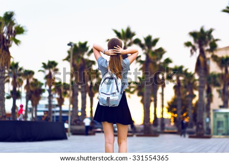 Outdoor lifestyle fashion portrait of young hipster woman walking at Barcelona, travel with backpack, stylish casual outfit, evening sunset, palms, student, blonde hairstyle, happy time, toned colors.
