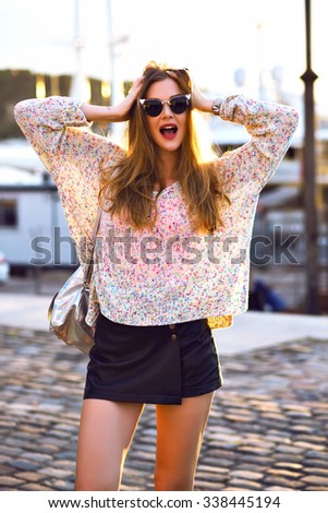 Positive outdoor portrait of stylish hipster blonde woman, casual clothes and sunglasses, positive surprised emotions, evening sunlight, warm colors.