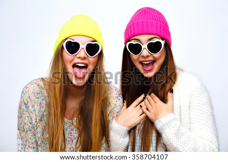 Trendy hipster portrait of young funny girls, hugs and having fun, going crazy together, party winter holiday time, neon sweaters hats vintage hearted sunglasses, emotions showing long tongue.