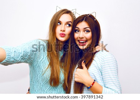 Positive friends portrait of two happy sister girls making selfie, sure funny faces, grimaces,joy, emotions, casual style, pastel colors, white wall. Sending air kiss, and say ok.