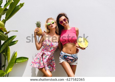Happy summer time of joyful amazing young women in attractive clothes with tropical fruits expressing to camera on white background outdoor. Sexy fashionable models having fun in summer vacation