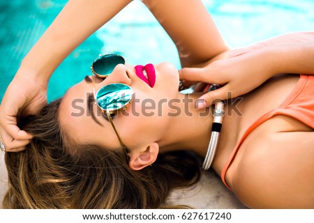 Close up portrait of sensual glamour model getting sunbathe near private pool, mirrored sunglasses, stylish jewelry, tanned skin, bright make up. Luxury vacation concept.