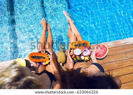 Summer pool party portrait of two best friends girls, sitting and holding plates with a lot of different tasty exotic fruits, health vegan lifestyle, relax, joy.