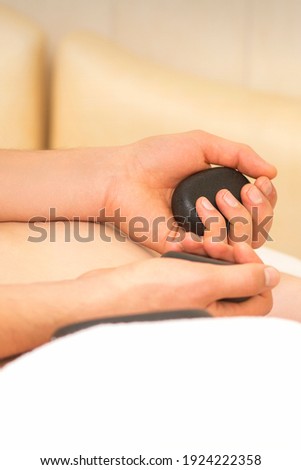 Hands of masseur hold black hot stones massaging back of a young adult woman in spa salon