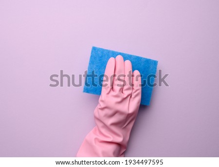 female hand in pink rubber glove holds blue kitchen sponge on purple background, close up