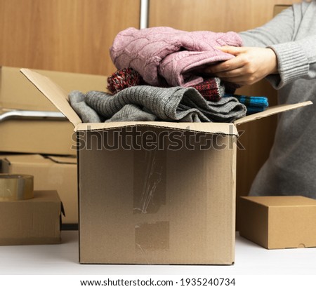 woman in a gray sweater collects clothes in a box, concept of assistance and volunteering, moving. Behind a stack of brown boxes