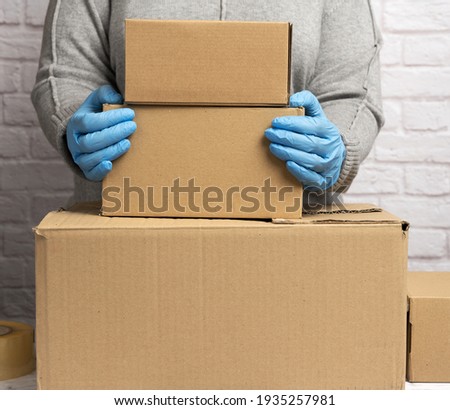 woman in a gray sweater is packing and blue gloves is holding a stack of brown cardboard boxes, moving, donation