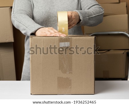 woman in a gray sweater holds a roll of duct tape and packs brown cardboard boxes on a white table, behind a stack of boxes. Moving concept