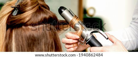 Back view of female hairdresser\'s hands curling women\'s hair with curling iron in a hair salon