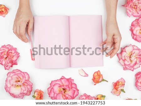 two female hands holding open notepad with clean pink sheets on a white background, around pink rosebuds, top view