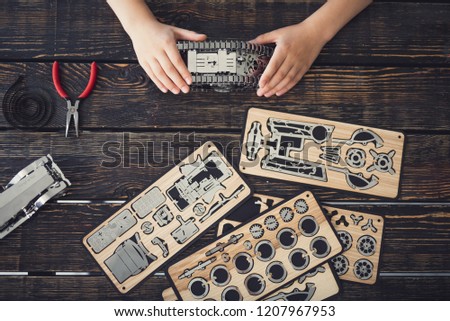 Top view of the table covered with pieces of wooden constructor and hands of child holding metal mechanism
