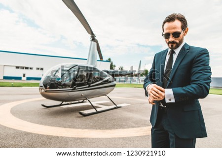 Checking the time. Thoughtful elegant businessman standing on the helicopter platform and looking at his watch