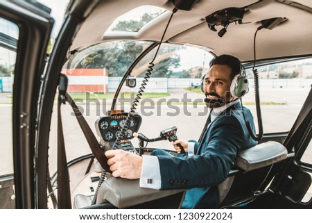 Cheerful bearded elegant man turning his head and smiling while sitting in the helicopter cabin and looking ready for the flight
