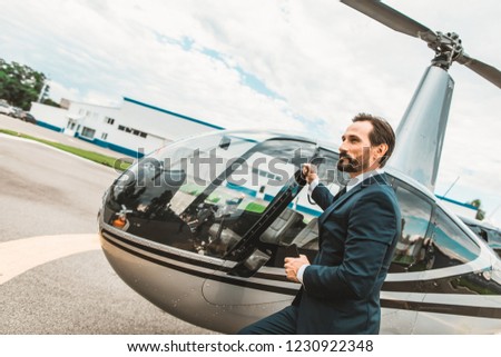 Handsome dark haired man in elegant suit standing near his personal helicopter and thoughtfully looking into the distance