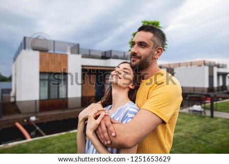 Joyful man is hugging his girlfriend and looking ahead with hint of smile on his face. Houses are on blurred background. Copy space on left side
