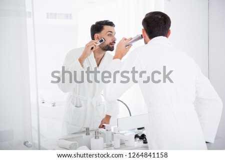 Concentrated young man in a white bathrobe looking at his reflection in the mirror while shaving his beard with a help of a shaving machine