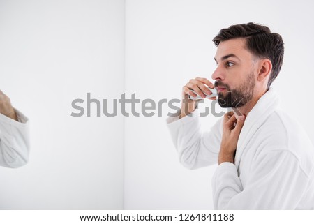 Calm attentive young man standing in front of the mirror and shaving beard while putting one hand on his neck