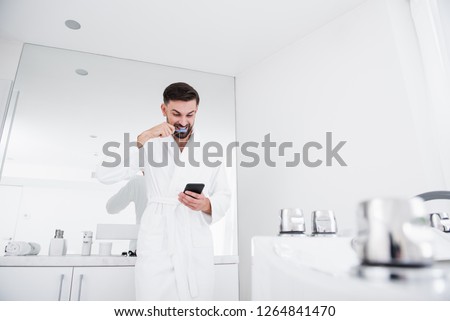 Emotional positive man standing in the bathroom in his bathrobe with a mirror behind and smiling. He looking at the screen of his modern gadget and brushing teeth