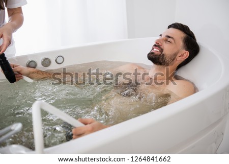 Relaxed bearded man smiling with his eyes closed while relaxing in the hydro massage bathtub of modern spa salon