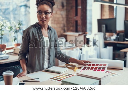 Waist up portrait of attractive young lady in glasses holding color palette while standing near table with cup of coffee, measuring tape and stationery. She is looking away with slight smile