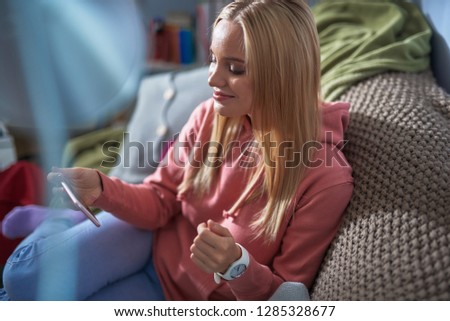 Side view portrait of charming blond girl sitting on couch and watching funny videos on smartphone. She looking at phone display and smiling