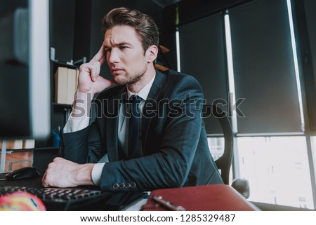 Calm elegant man in dark sit sitting at his workplace and frowning while thoughtfully looking at the screen of his computer