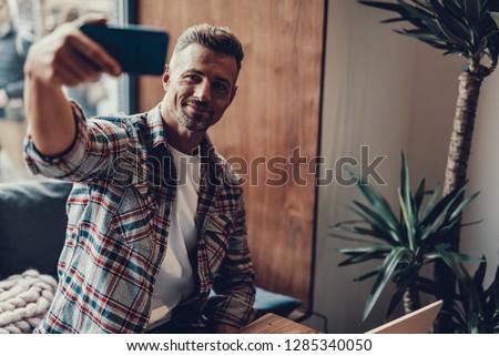 Peaseful and unshaven mature man in street style clothes sitting inside comfort cafe and making selfie on cellular
