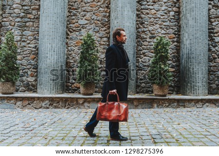 Full length profile portrait of stylish man standing in old street in city. He is wearing classic clothes and carrying handbag. Copy space in left side