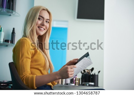 Portrait of attractive blond girl in yellow sweater holding brochure with list of services. She looking at camera and smiling