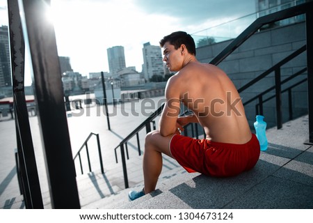 Side view serene bare-chested man resting while sitting on stairs after good training outdoor. Bottle of water locating opposite him