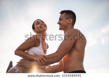 Waist up of the attractive young man standing shirtless and smiling while putting his hands on the waist of beloved woman