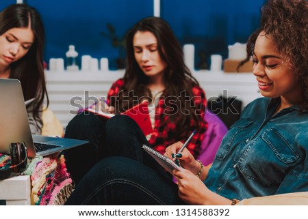 Focus on outgoing woman writing in notebook. She leaning on chair in room. Her friends sitting on background
