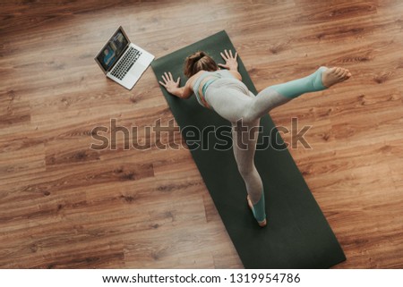 Health practice concept. Top view portrait of young balanced woman in sportswear doing one legged dolphin yoga pose on mat at home with notebook beside