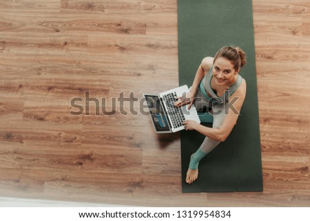 Technology in our life. Full length top view portrait of young smiling lady after yoga practice sitting on mat with notebook and looking up on camera. Copy space on left