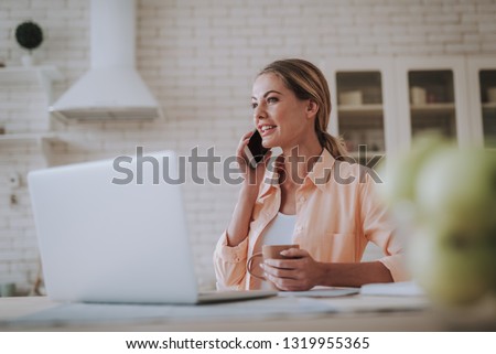 Pleased young woman sitting in the kitchen with a cup of tea and smiling while talking on the phone