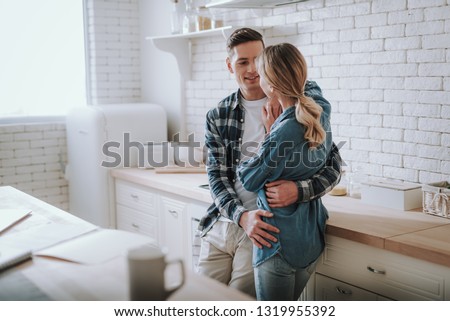 Waist up of the couple in casual clothes looking cute while hugging in the white kitchen