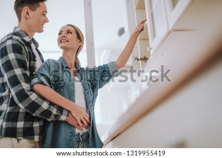Happy young woman putting one hand on the shelf of cupboard and smiling while turning to her beloved man