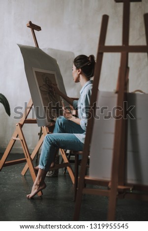 Dark haired lady in casual clothes sitting barefooted in her art studio and drawing a person on the canvas
