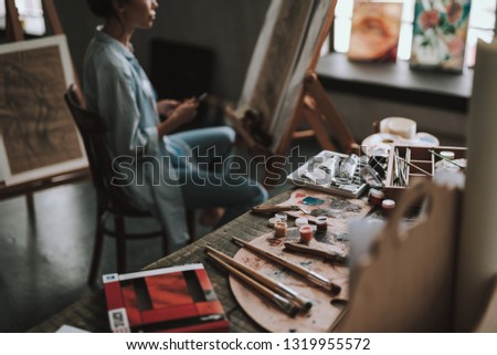 Selective focus of the painting equipment on the table in art studio and female artist sitting on the background