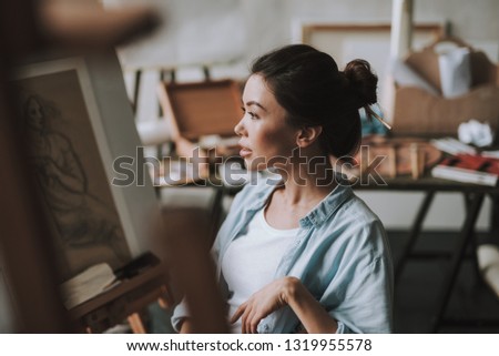 Young beautiful female artist sitting in art studio with art supplies on the background and thoughtfully looking into the distance