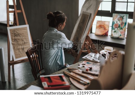 Young female artist sitting at the table with creative tools and turning to the drawing on the easel