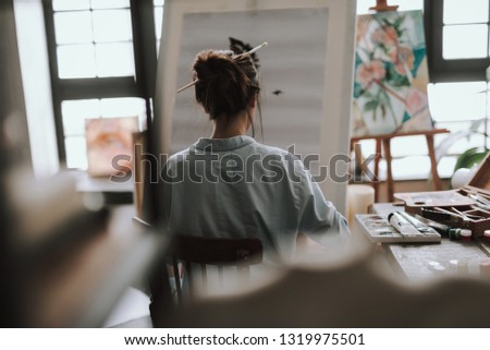 Dark haired female artist sitting in the art studio in front of the canvas on the easel with art supplies on the table