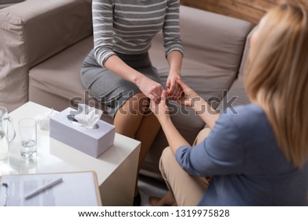 A female psychologist trying to calm down patient. They are sitting on the sofa in the office and keeping hands together. Focus on arms